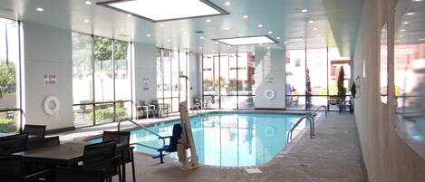 Un área de piscina interior con paredes de vidrio transparente, una escalera de piscina y mesas y sillas alrededor.