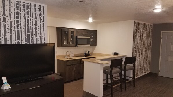 Cocina compacta con televisor, microondas y pileta. Hay dos taburetes de bar y una pequeña isla.