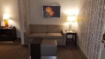 Een hotelkamer met een beige bank, een klein tafeltje met een lamp, een nachtkastje met een lamp, een decoratief wandobject en een kamerbreed tapijt met dessin.
