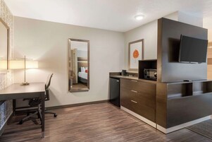 Una habitación de hotel moderna con suelo de madera, un escritorio con lámpara, un espejo grande, un armario, un televisor y una pequeña cocina.
