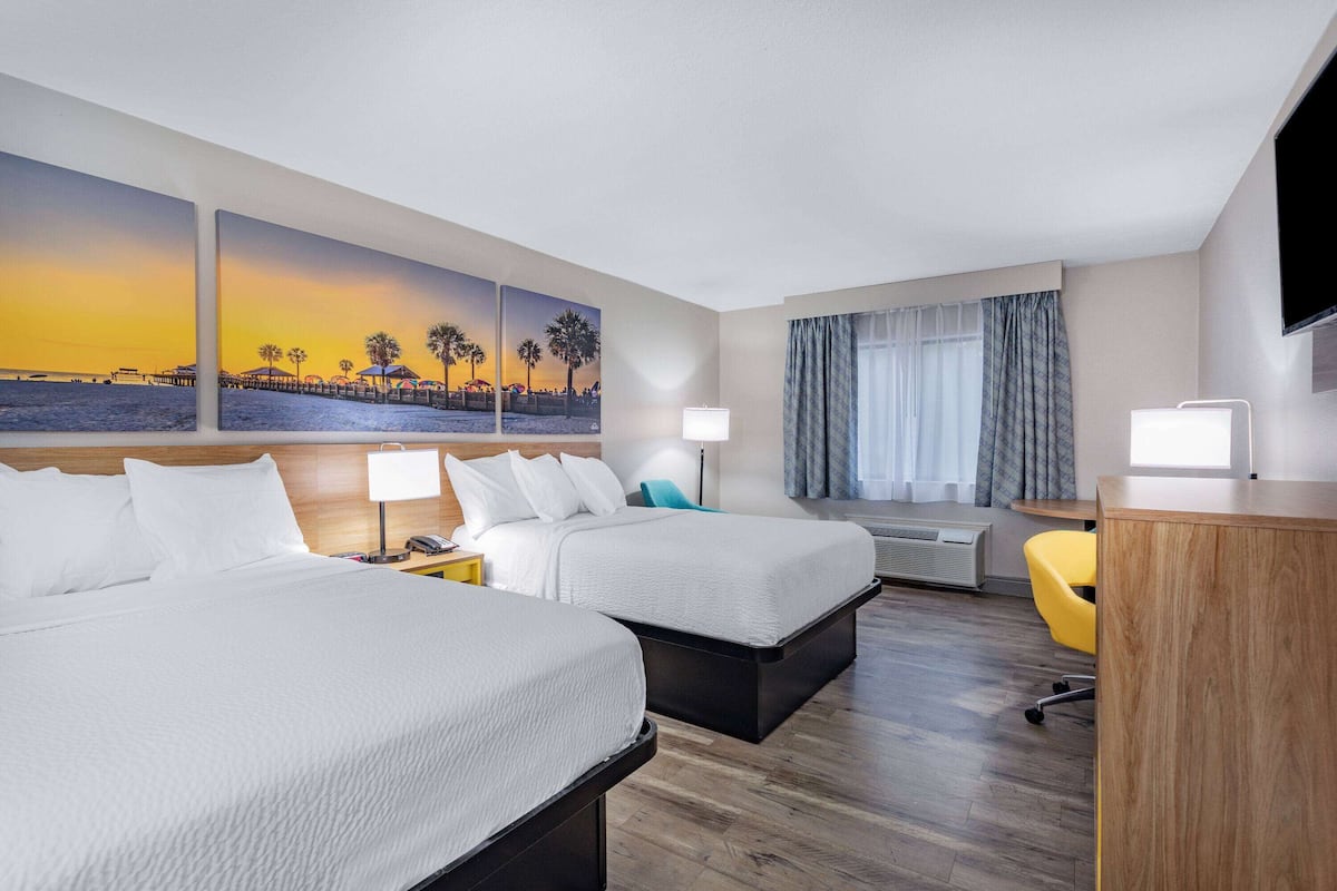Ein Hotelzimmer mit zwei Betten, einem Schreibtisch, einem Stuhl, einem Flachbildfernseher, einem Fenster mit Vorhängen und einem Wandbild.