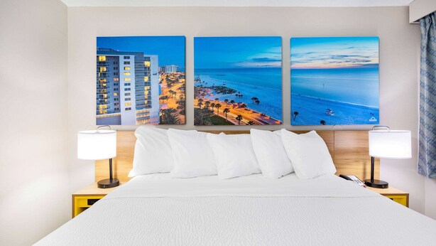 Habitación de hotel con una cama grande, ropa blanca de cama, dos lámparas de noche y tres cuadros enmarcados con motivos costeros en la pared.