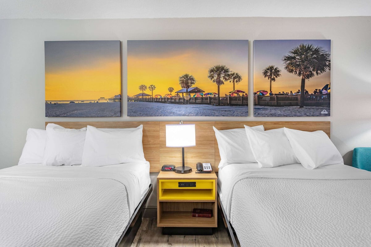 Ein Hotelzimmer mit zwei Betten, einem Nachttisch mit Lampe, einer digitalen Uhr und einem kleinen Tisch.