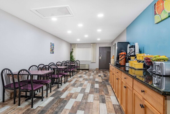 A dining area with wooden chairs and tables, a kitchen counter with appliances, and a blue wall with a fruit artwork.