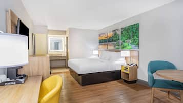 Habitación de hotel con una cama, un escritorio con lámpara, una silla, una mesita y un cuadro en la pared.