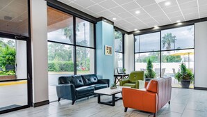 Un vestíbulo moderno con un sofá de cuero negro, un sillón naranja y una silla verde. Hay ventanas amplias con vistas a zonas verdes y una palmera.