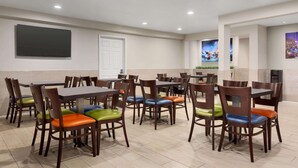A dining area with multiple tables and chairs, a flat-screen TV mounted on the wall, and a door in the background. - Days Inn & Suites by Wyndham Anaheim At Disneyland Park (Anaheim)
