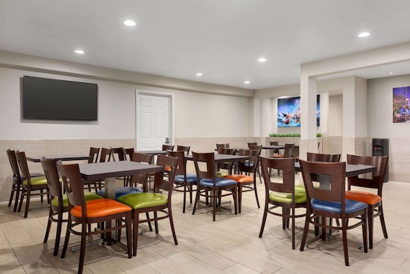 Un espace repas avec plusieurs tables et chaises, un téléviseur à écran plat fixé au mur et une porte visible au fond.