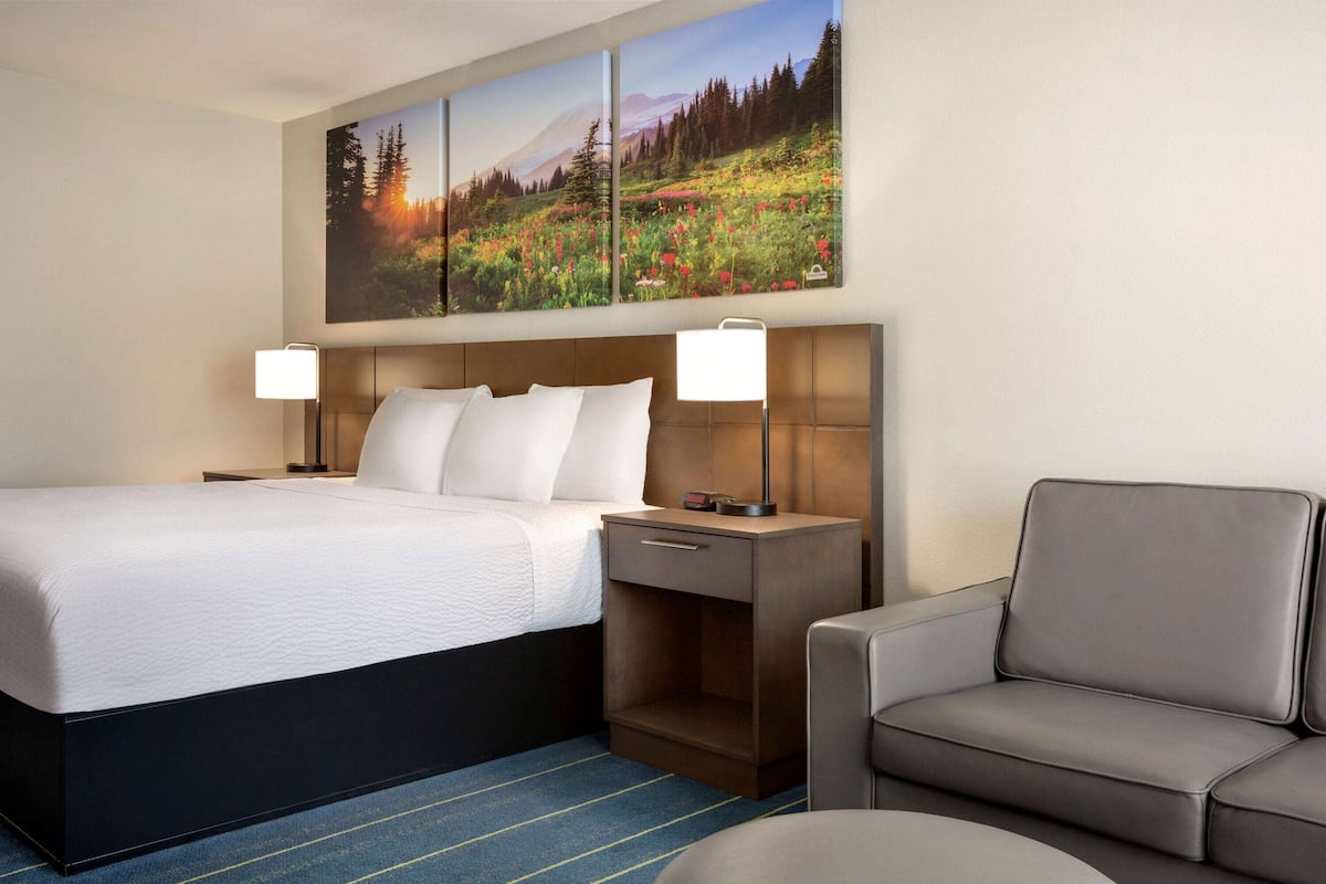 A hotel room with a bed, two bedside lamps, a nightstand, a sofa, and a large landscape painting on the wall.