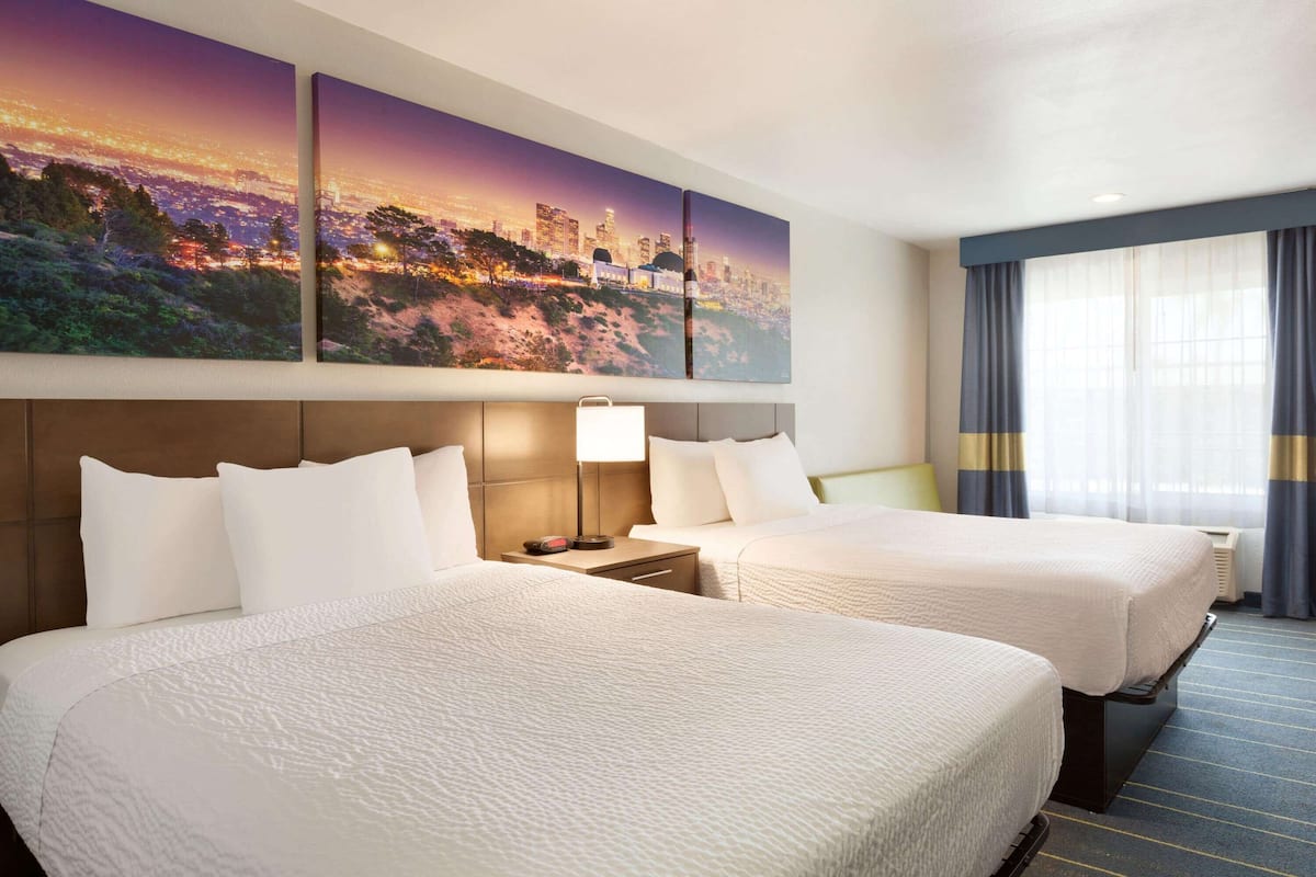 A hotel room with two beds, a nightstand, a lamp, and a large cityscape painting on the wall.