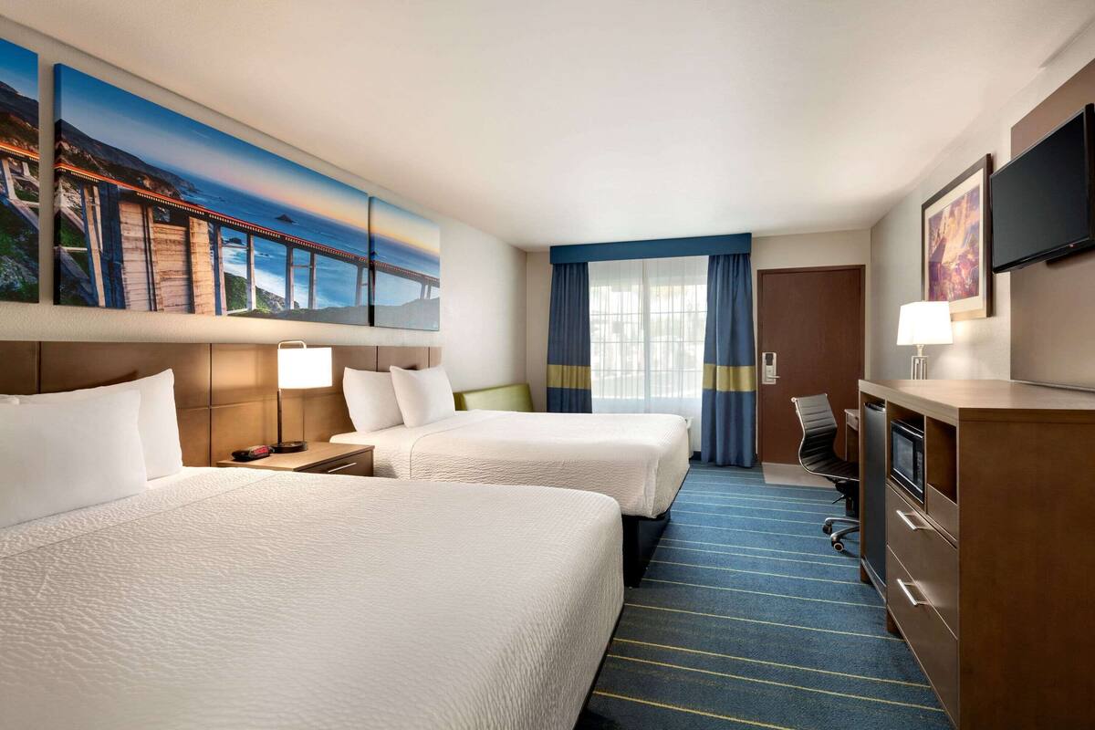 A hotel room with two beds, a desk, a television, and a large window with curtains.