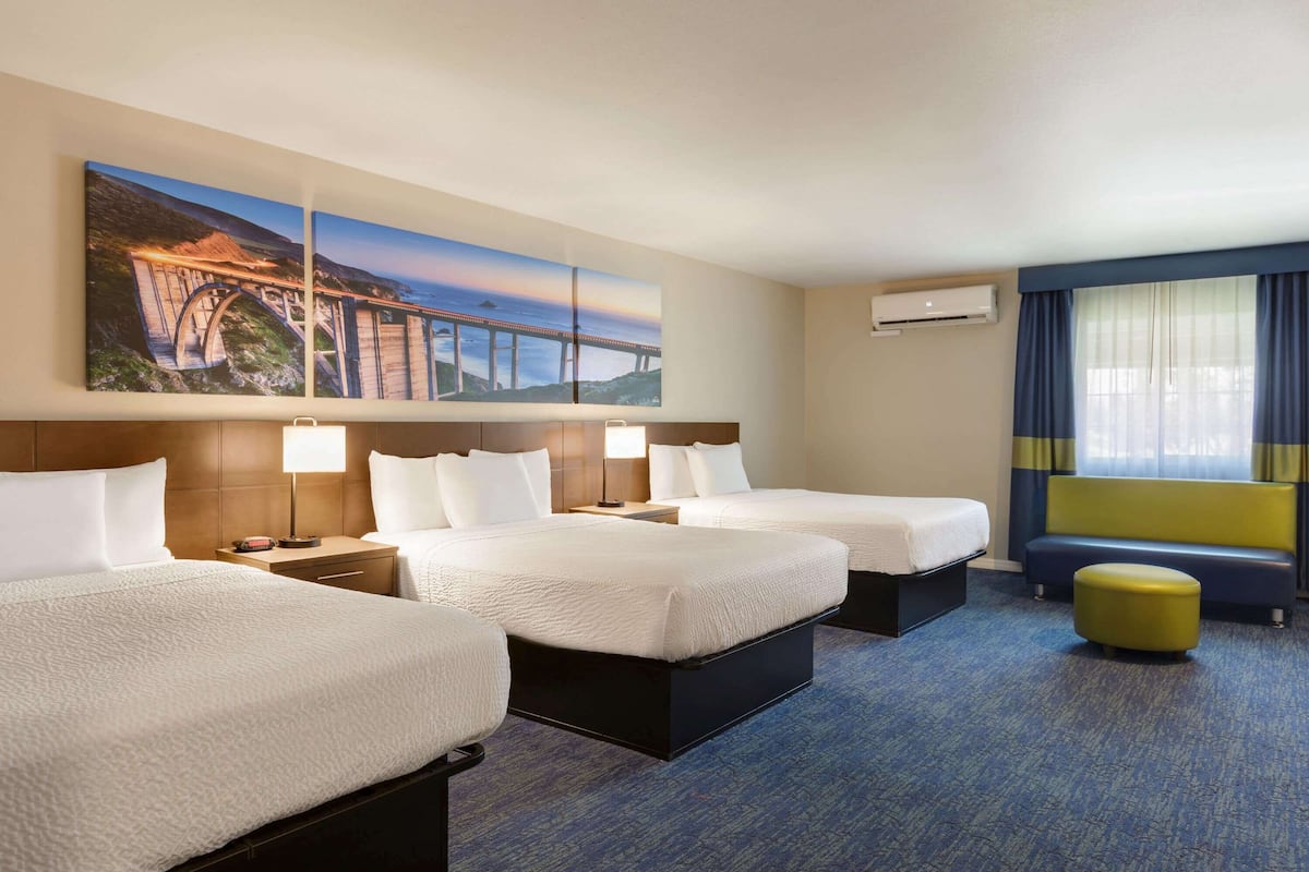 A hotel room with two beds, a bench, a large window with curtains, and a painting of a bridge.