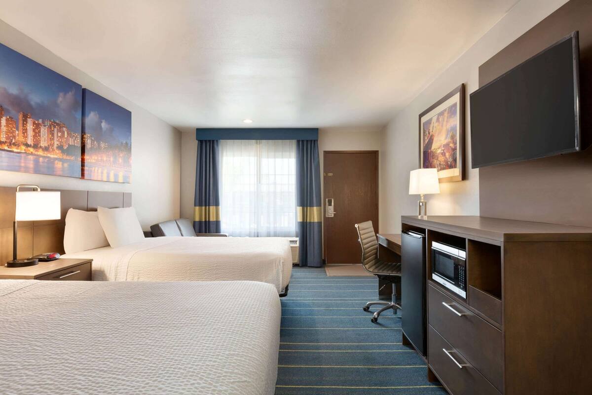 A hotel room with two beds, a desk, a TV, and a cityscape painting.