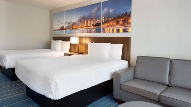 A hotel room with two beds, a sofa, and a cityscape painting on the wall. - Days Inn & Suites by Wyndham Anaheim At Disneyland Park (Anaheim)