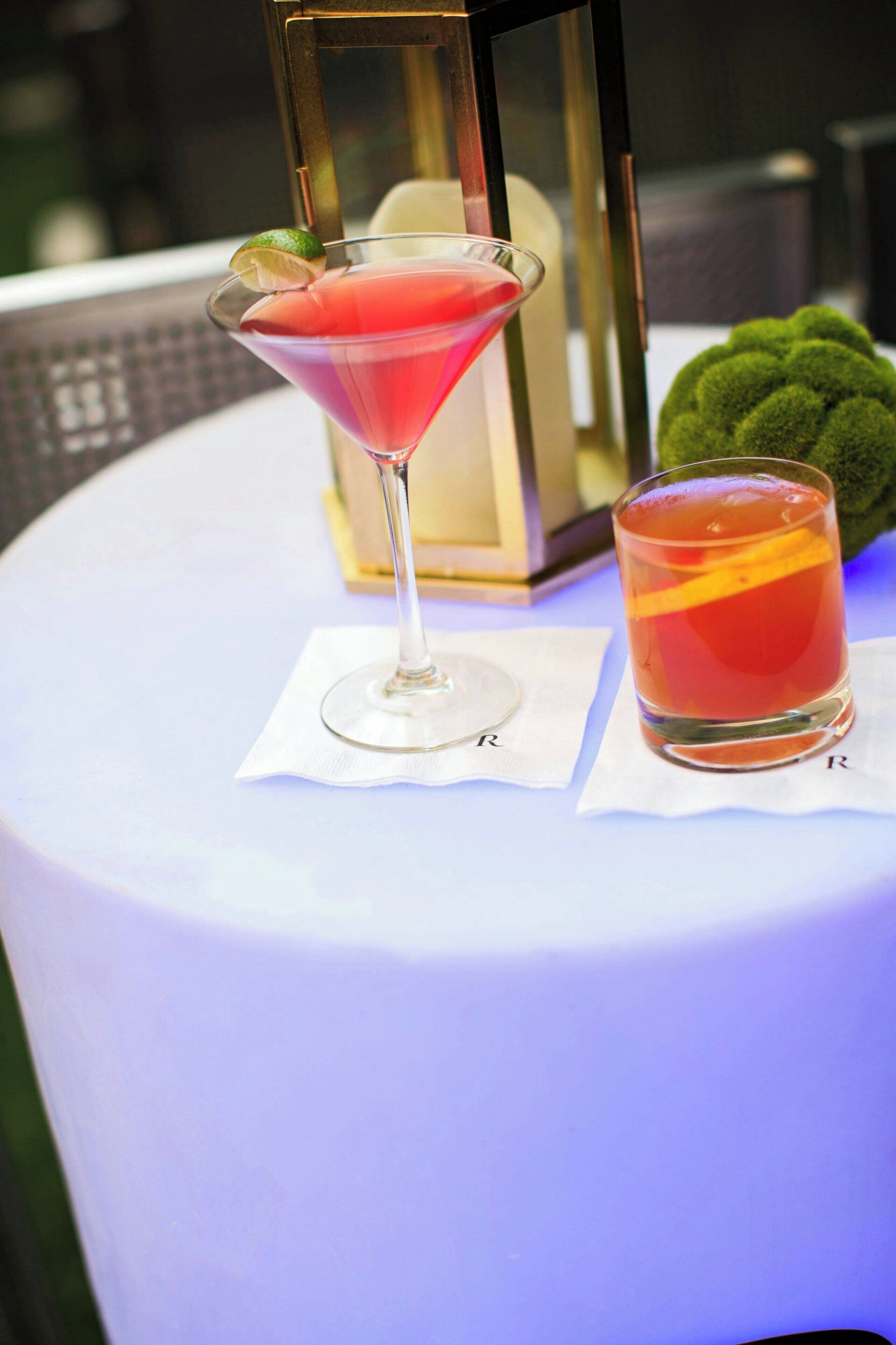 A martini glass with a lime garnish on a white tablecloth, accompanied by a cocktail glass with an orange slice.