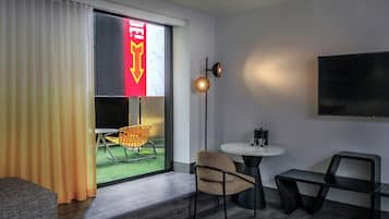 Quarto de hotel moderno com uma grande janela, uma TV de tela plana na parede, uma pequena mesa redonda, uma cadeira, um sofá e uma escrivaninha com cadeira.