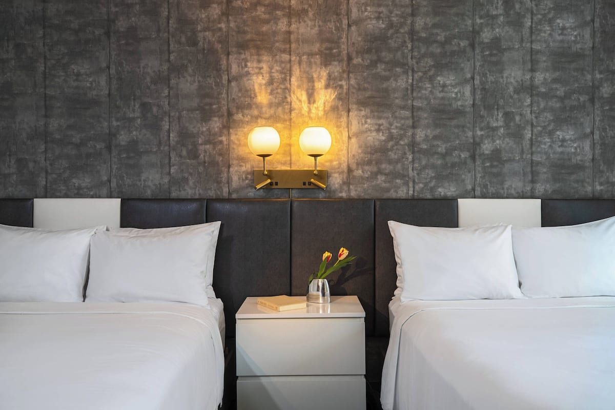 Una habitación de hotel con dos camas, una mesita de noche con un jarrón de flores e iluminación en la pared.