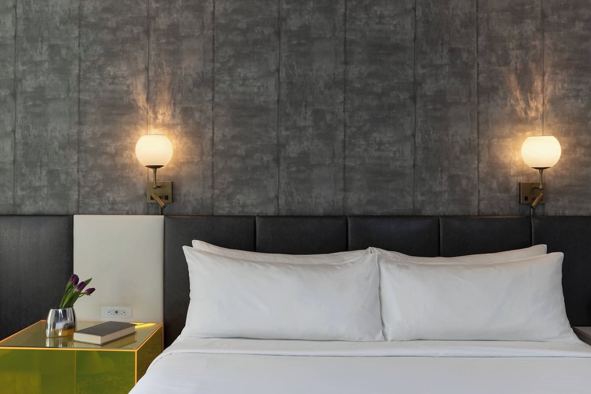 Habitación de hotel moderna con una cama grande, dos lámparas de noche, una mesita de noche con un libro y un jarrón de flores, y una pared texturizada detrás de la cama.