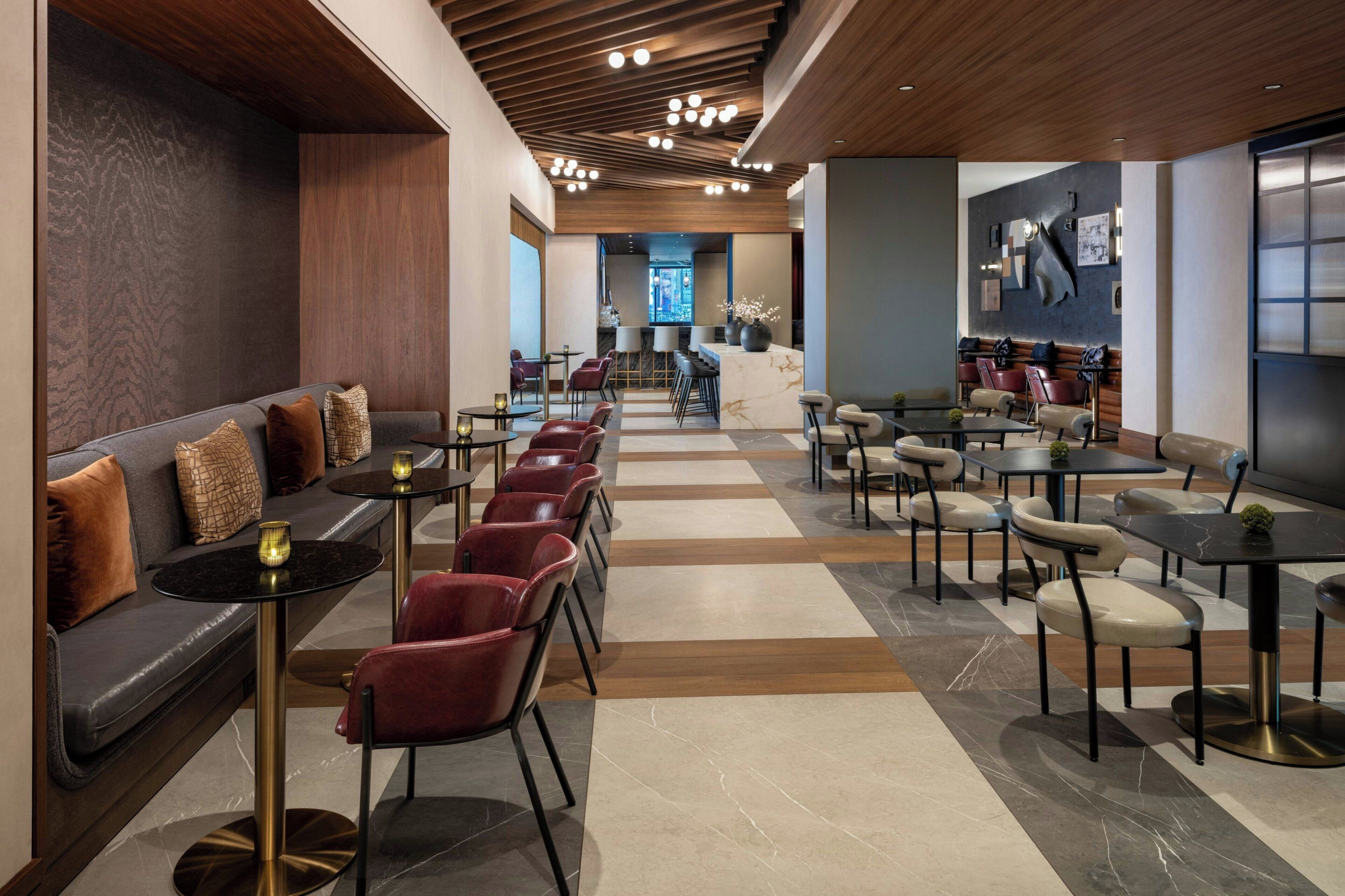 Een modern interieur met een lange rij rode stoelen en tafels, een zithoek met een bank, en een bar met een marmeren achterwand.