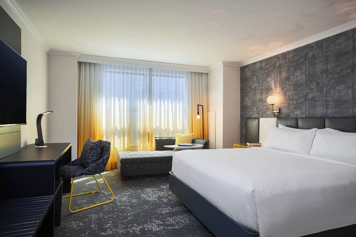 Habitación de hotel con una cama grande, un escritorio con silla, un pequeño rincón de descanso y un amplio ventanal con cortinas.