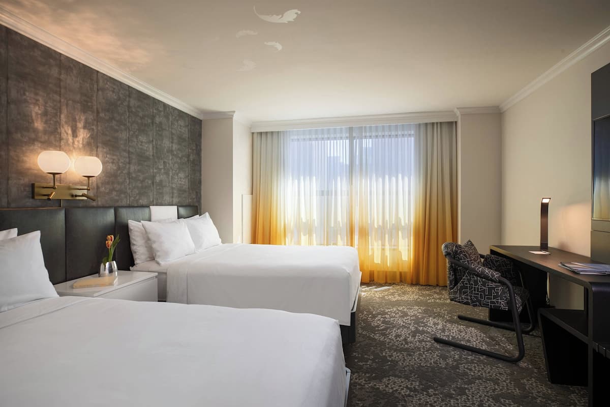 Habitación de hotel con dos camas, un escritorio, una silla y un amplio ventanal con cortinas.