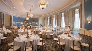 A grand ballroom with round tables set for a formal event, featuring chandeliers and elegant chairs.