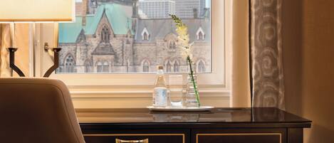 A hotel room with a desk, a lamp, a vase with flowers, and a view of a historic building.