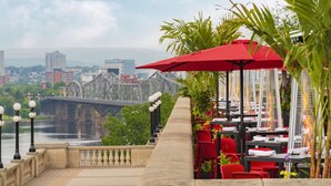 Un área de comedor al aire libre con sillas y mesas rojas, un paraguas rojo y vista a un puente y al paisaje urbano de fondo.