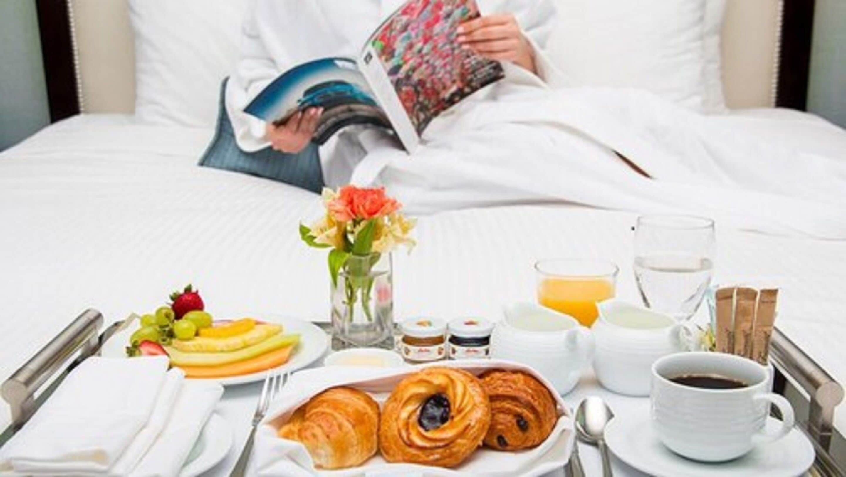 A neatly arranged breakfast table with croissants, fruits, and beverages, set on a bed with a person reading a magazine.