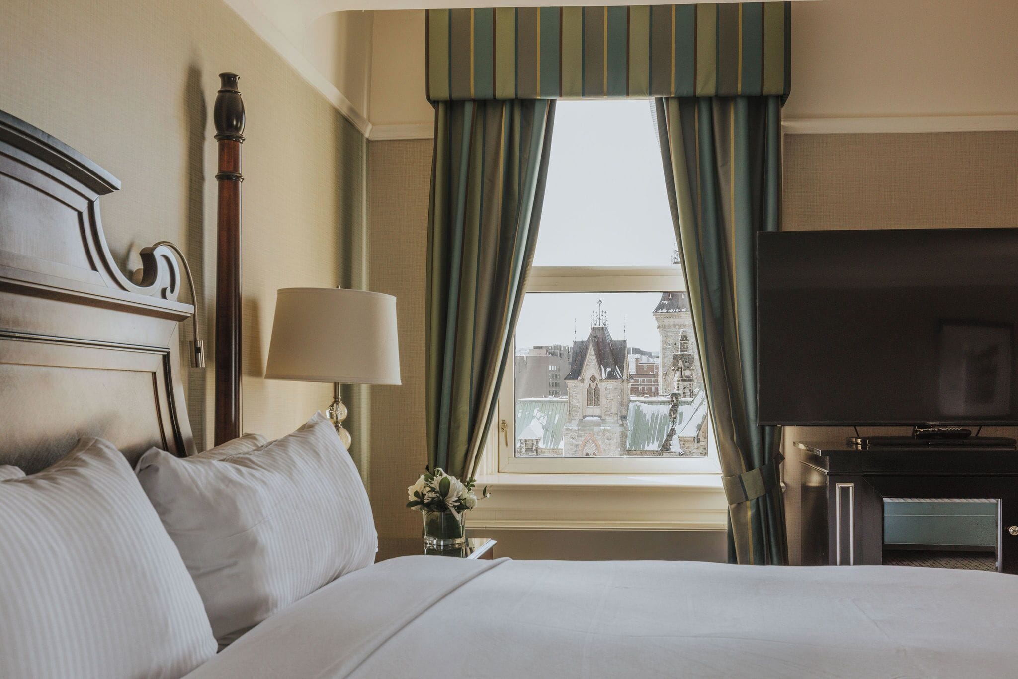 A hotel room with a large bed, a television, a window with curtains, and a view of a cityscape.