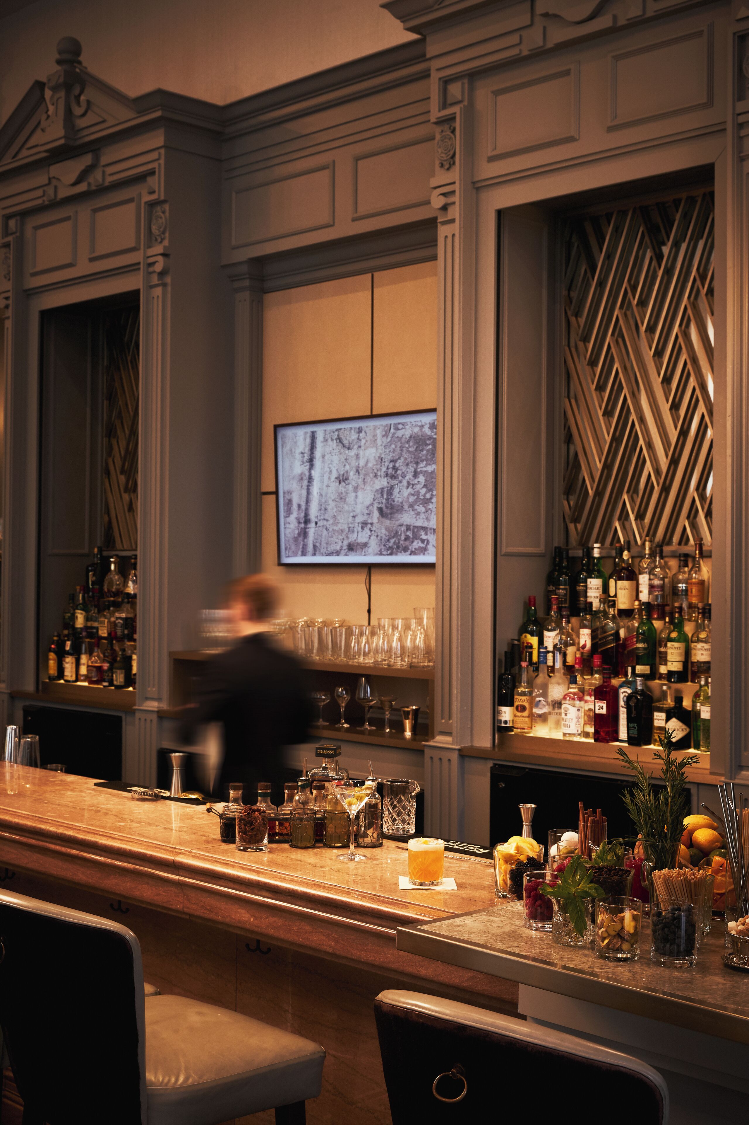 A bar area with a long counter, shelves of bottles, glasses, and a television screen.