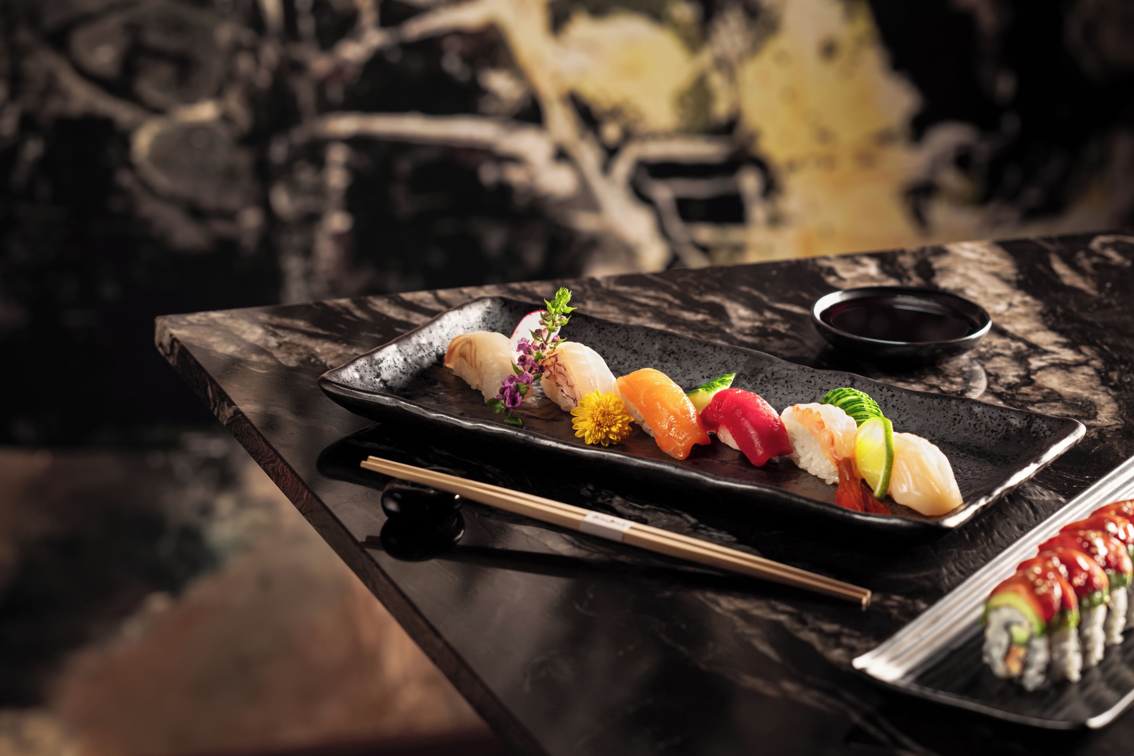 A sushi platter on a black slate plate with chopsticks and a small dish of soy sauce.