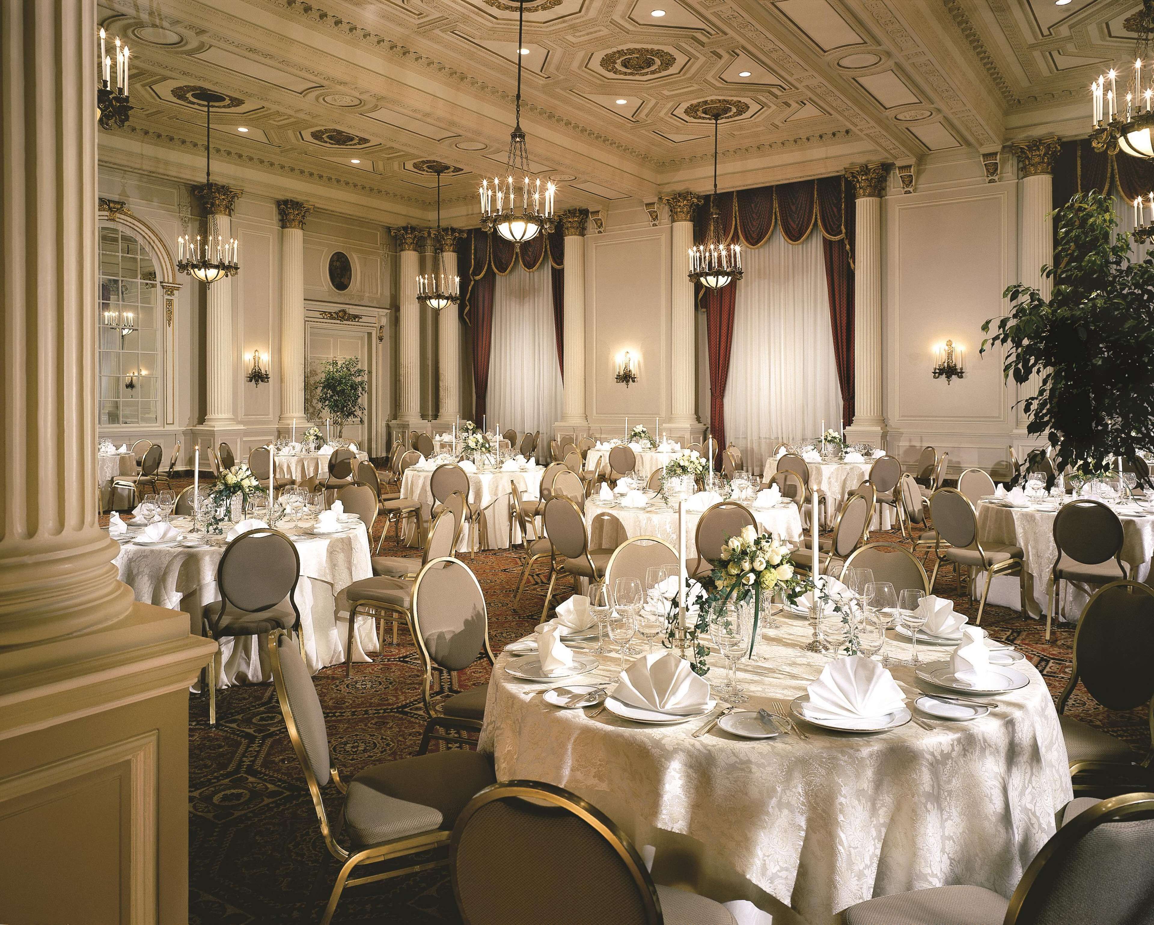 A banquet hall with round tables set for a formal event, featuring chandeliers, draped curtains, and a classic architectural ceiling.