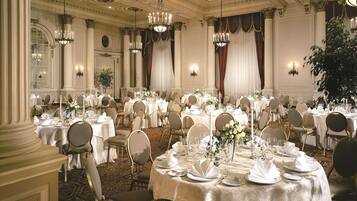 A banquet hall with round tables set for a formal event, featuring chandeliers, draped curtains, and a classic architectural ceiling.