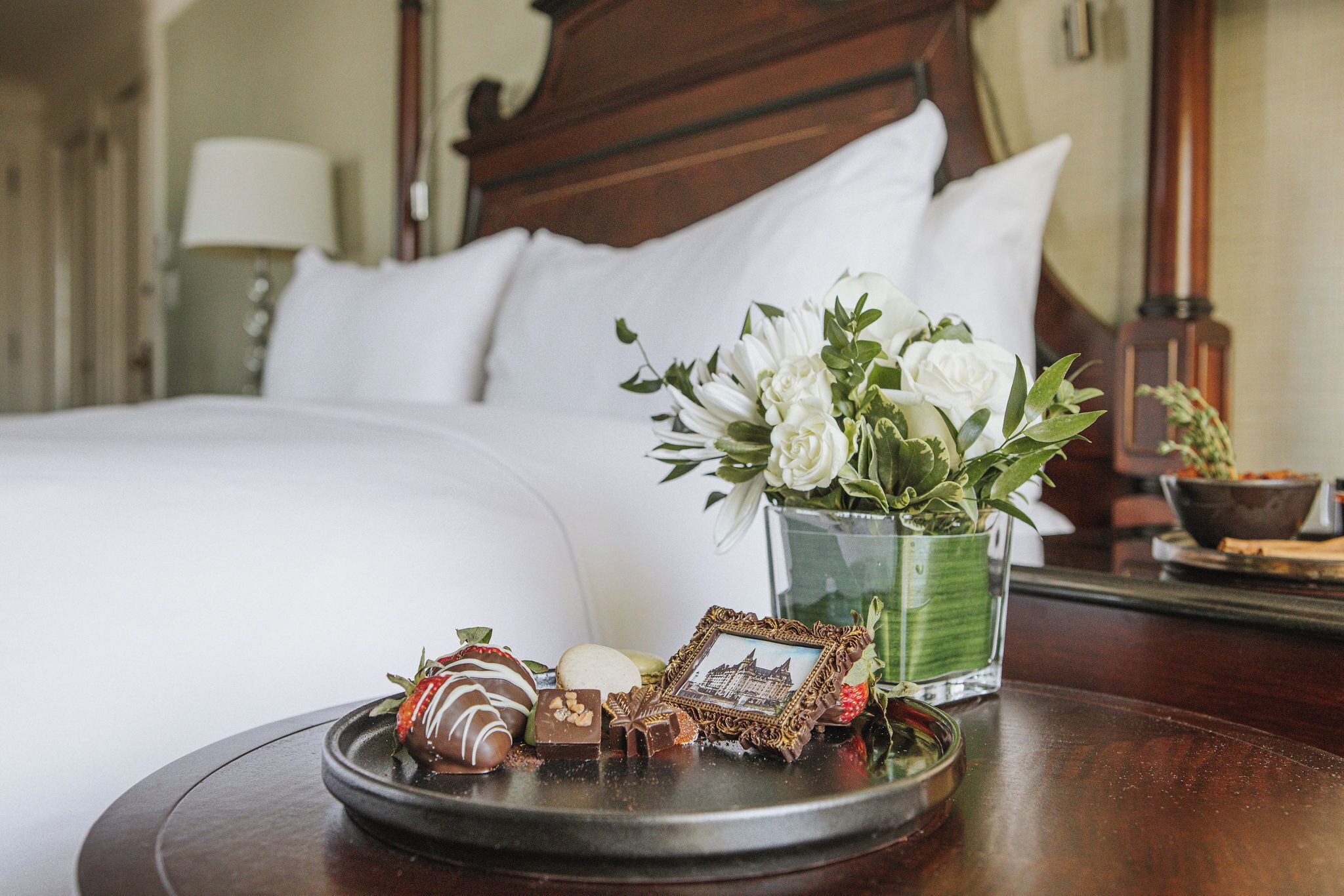 Een netjes opgemaakt bed met witte lakens, een nachtkastje met een glazen vaas vol bloemen, en een dienblad met diverse chocolades en een klein fotolijstje.