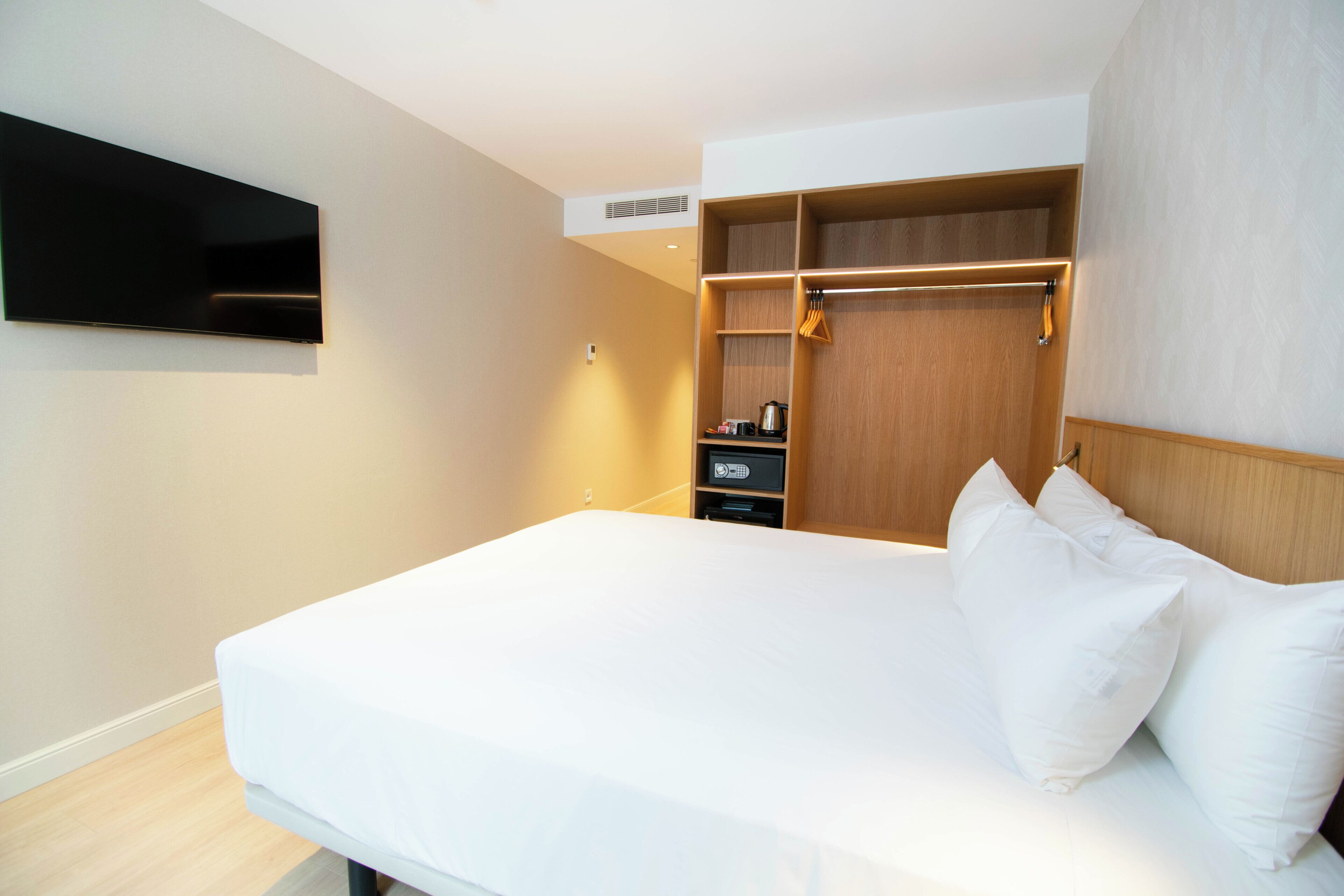 A hotel room with a bed, a TV mounted on the wall, a wardrobe, and a shelf with a kettle.