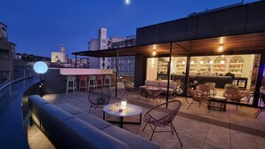 Un área de estar al aire libre con bar, asientos acolchados y vistas a la ciudad al anochecer.