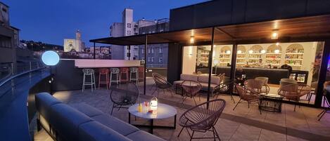 An outdoor seating area with a bar, cushioned seating, and a view of the cityscape at dusk.
