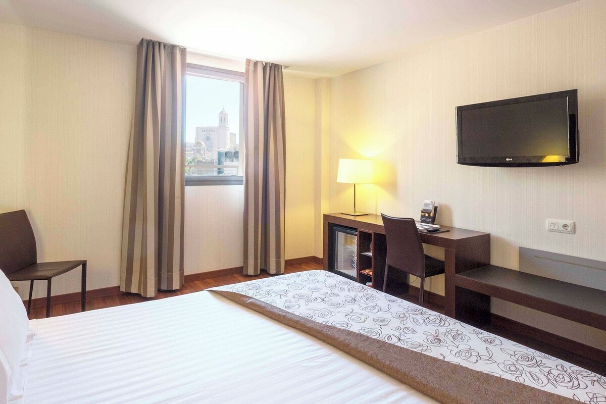 A hotel room with a bed, a desk, a chair, a television, and a window with curtains.