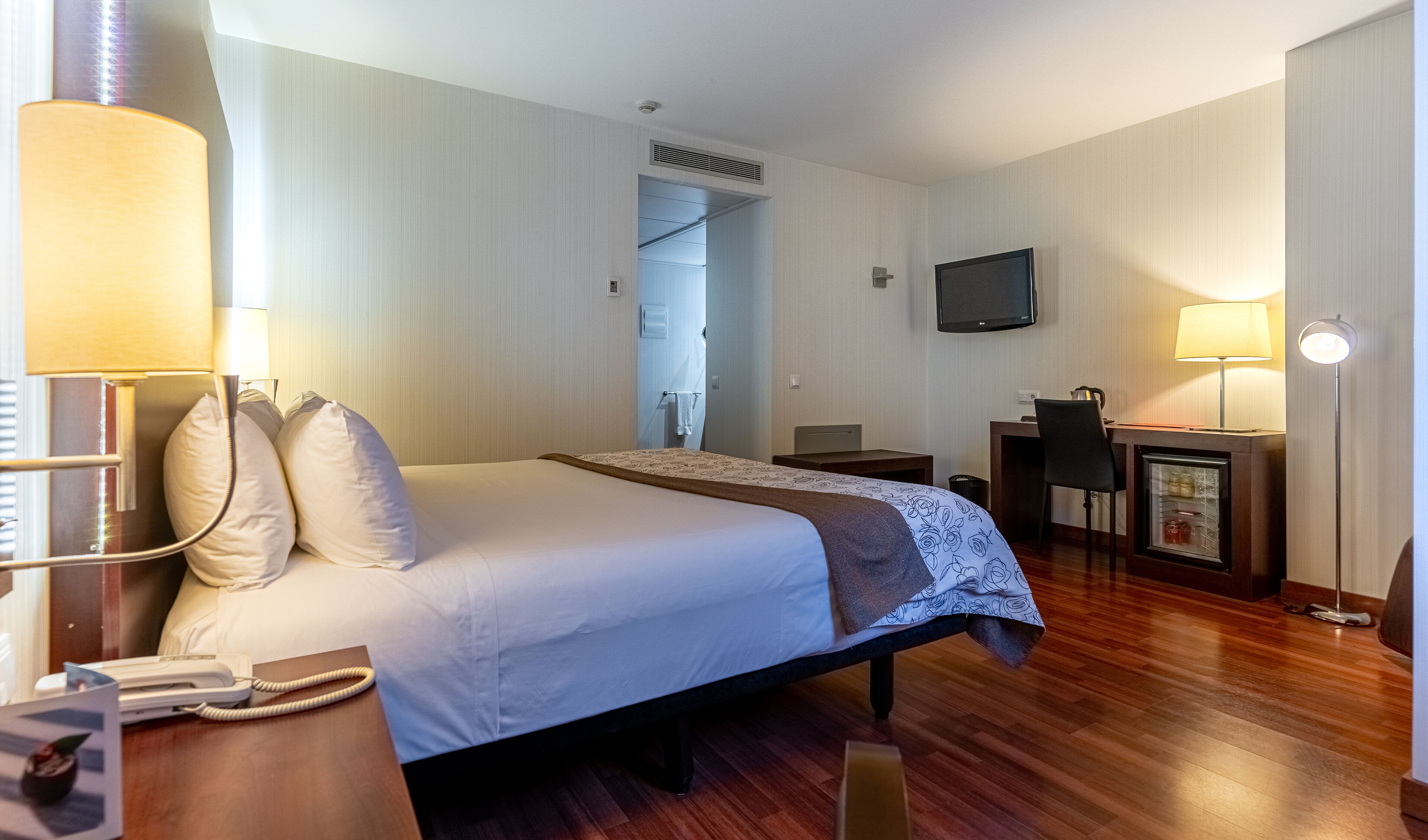 A hotel room with a bed, bedside table with a lamp, a desk with a chair, a television, and a door leading to a bathroom.