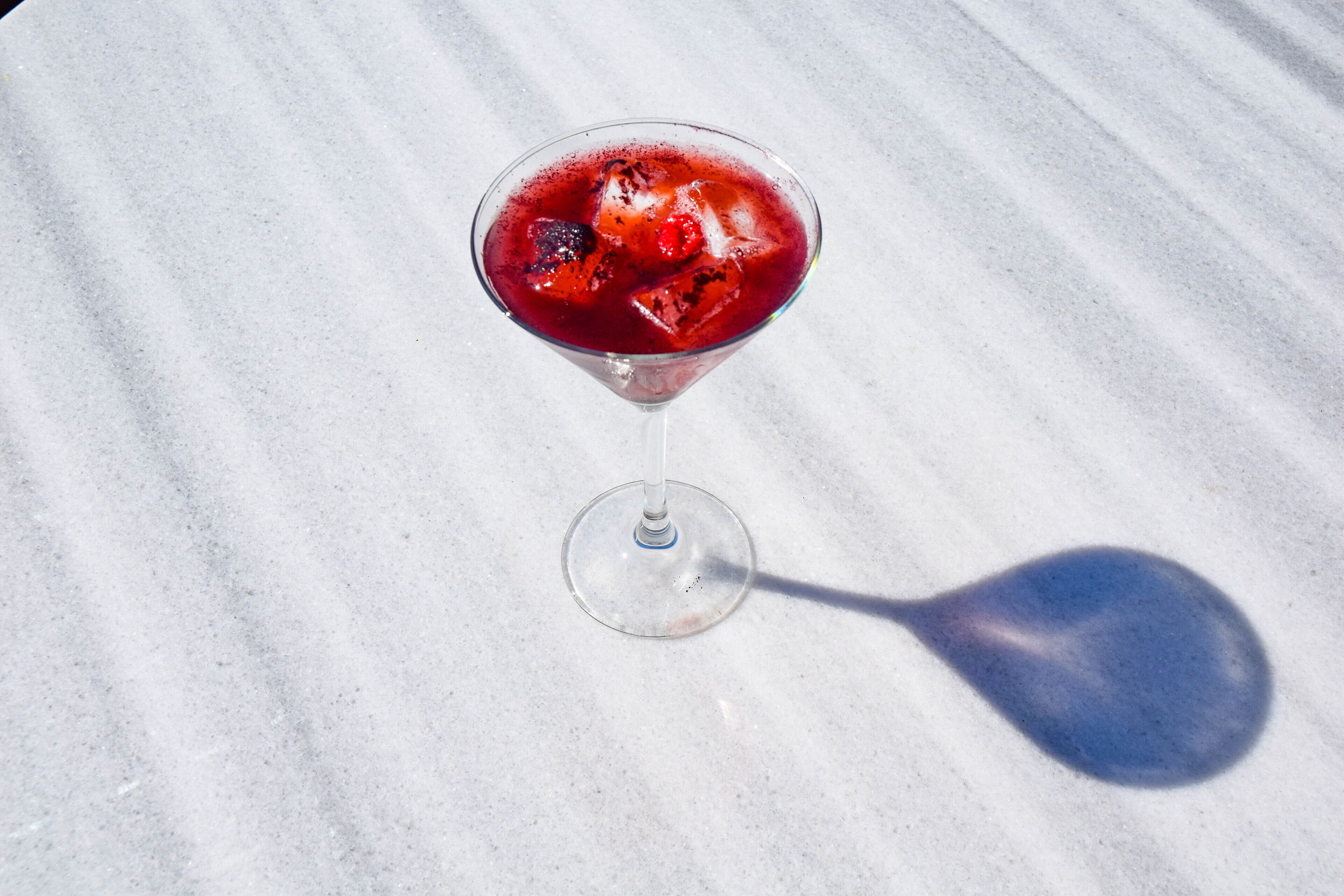Una copa de martini con una bebida roja y cubitos de hielo sobre una superficie de mármol.