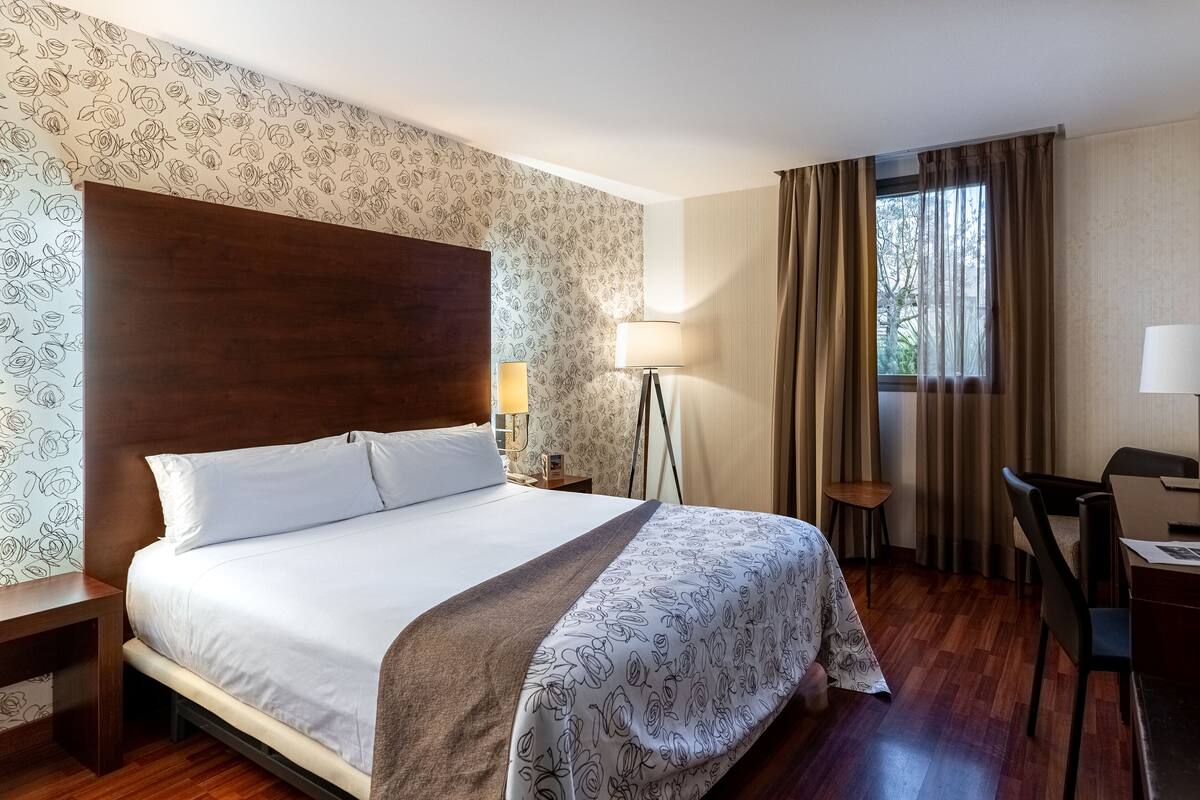 Ein Hotelzimmer mit einem großen Bett, einem Schreibtisch, einem Stuhl, einer Lampe und einem Fenster mit Vorhängen.