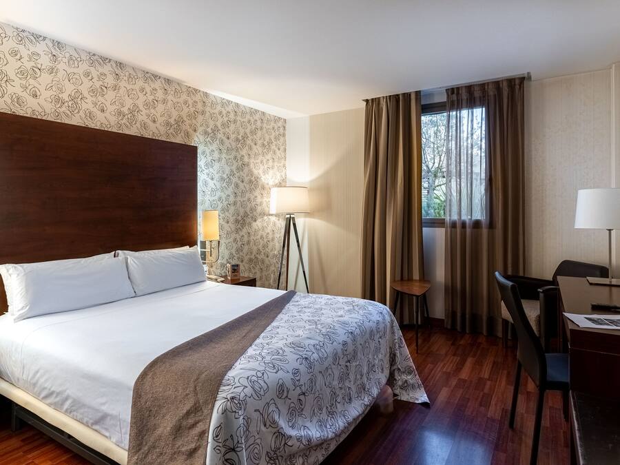 A hotel room with a large bed, a desk, a chair, a lamp, and a window with curtains.