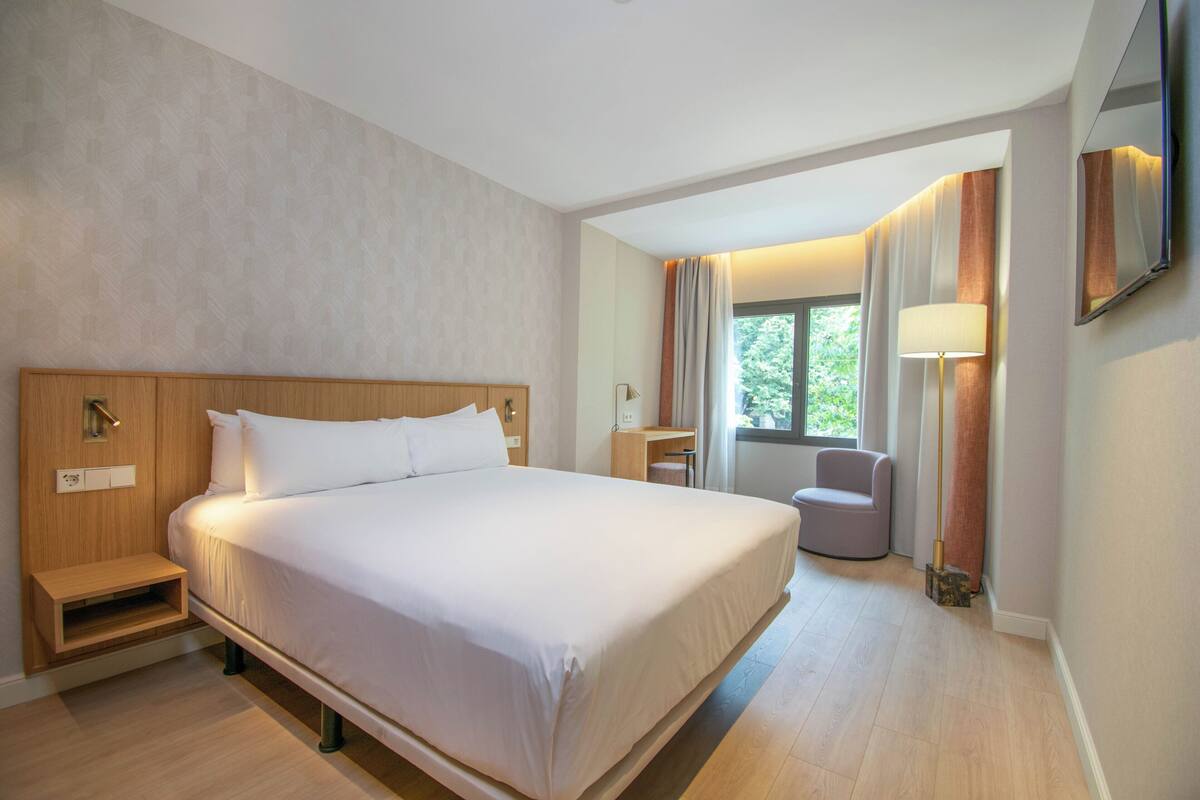 Ein Hotelzimmer mit einem großen Bett, Nachttischen, einem Schreibtisch, einem Stuhl und einem Fenster mit Vorhängen.