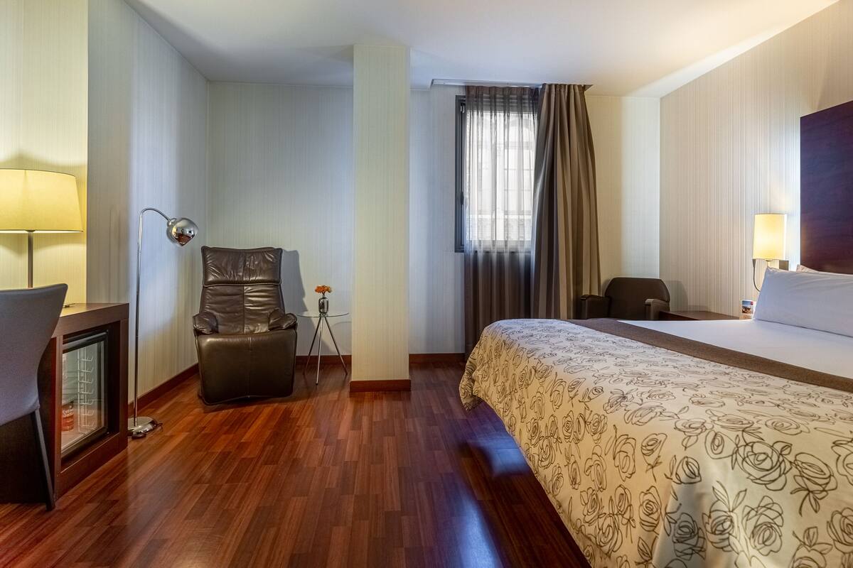 Ein Hotelzimmer mit einem Bett, einem Schreibtisch, einem Stuhl, einer Lampe und einem Fenster mit Vorhängen.