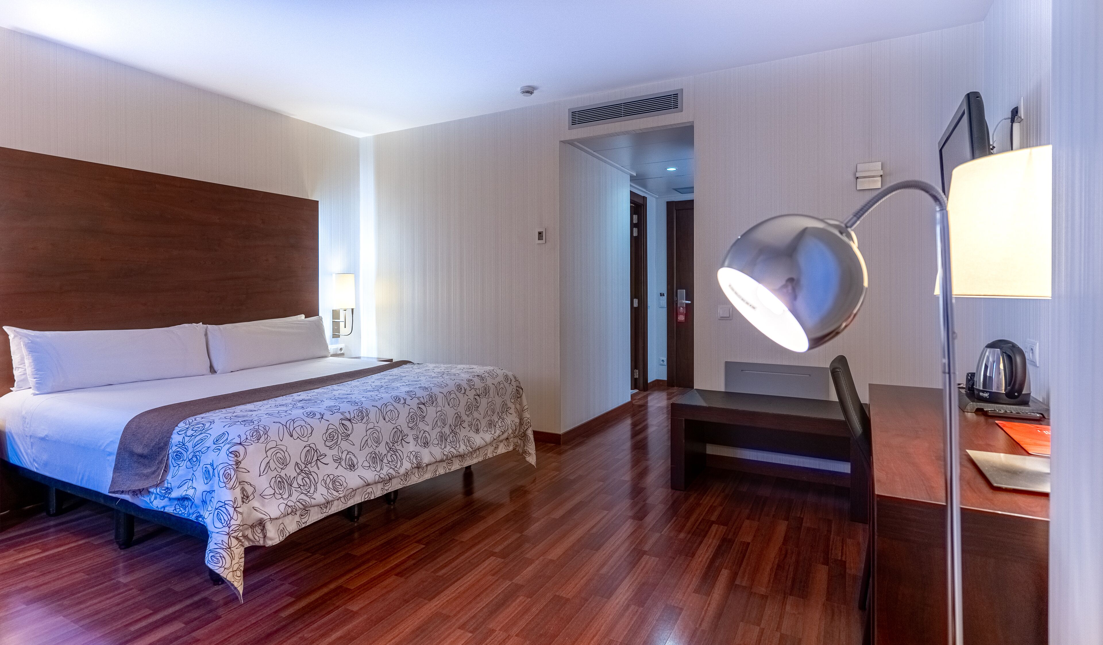 Ein Hotelzimmer mit Holzboden, einem Bett mit floralem Überwurf, einem Schreibtisch mit Lampe und Blick auf den Flur.