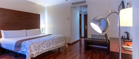 A hotel room with a wooden floor, a bed with a floral bedspread, a desk with a lamp, and a view of the hallway.