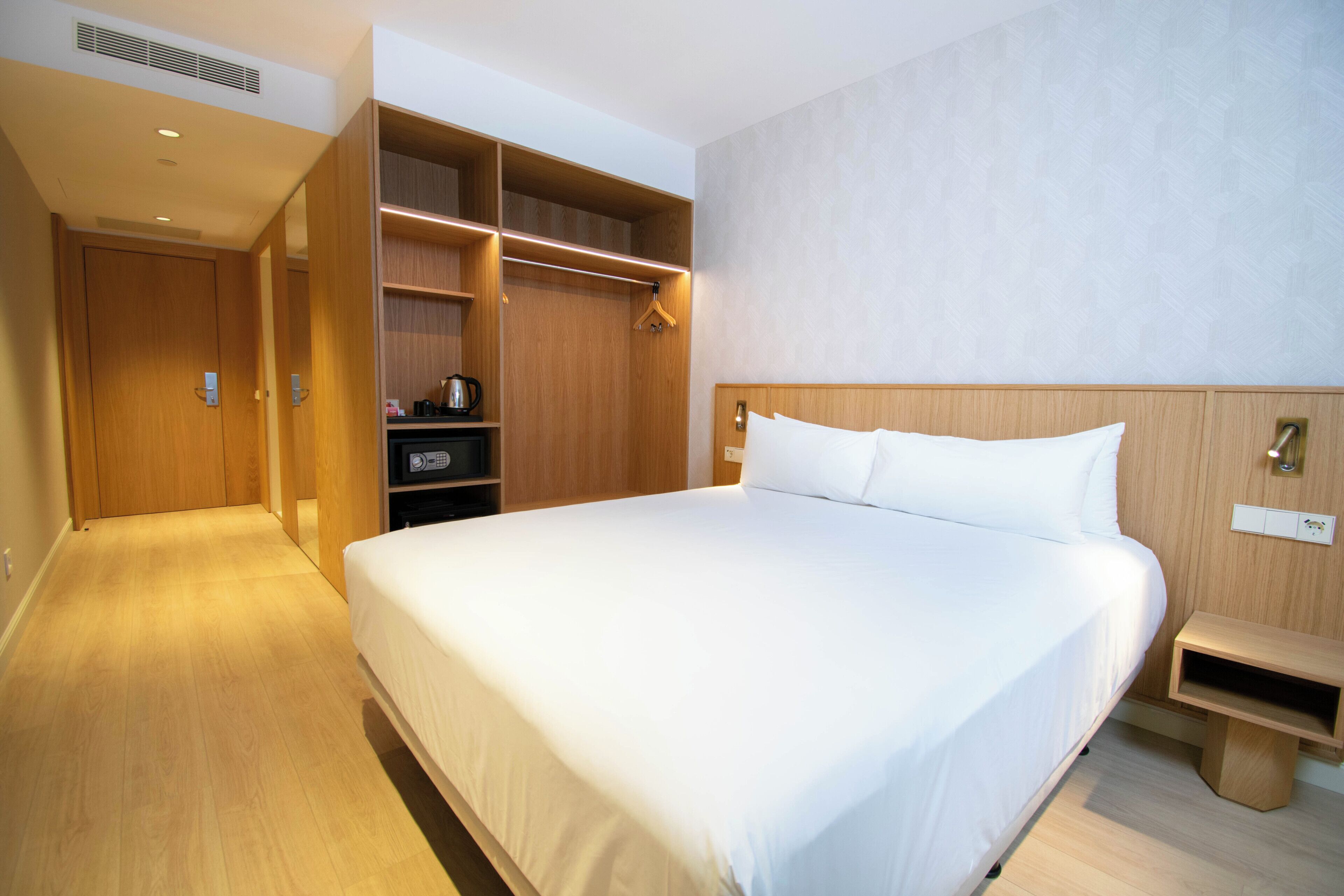 A hotel room with a large bed, wooden wardrobe, and a bedside table.
