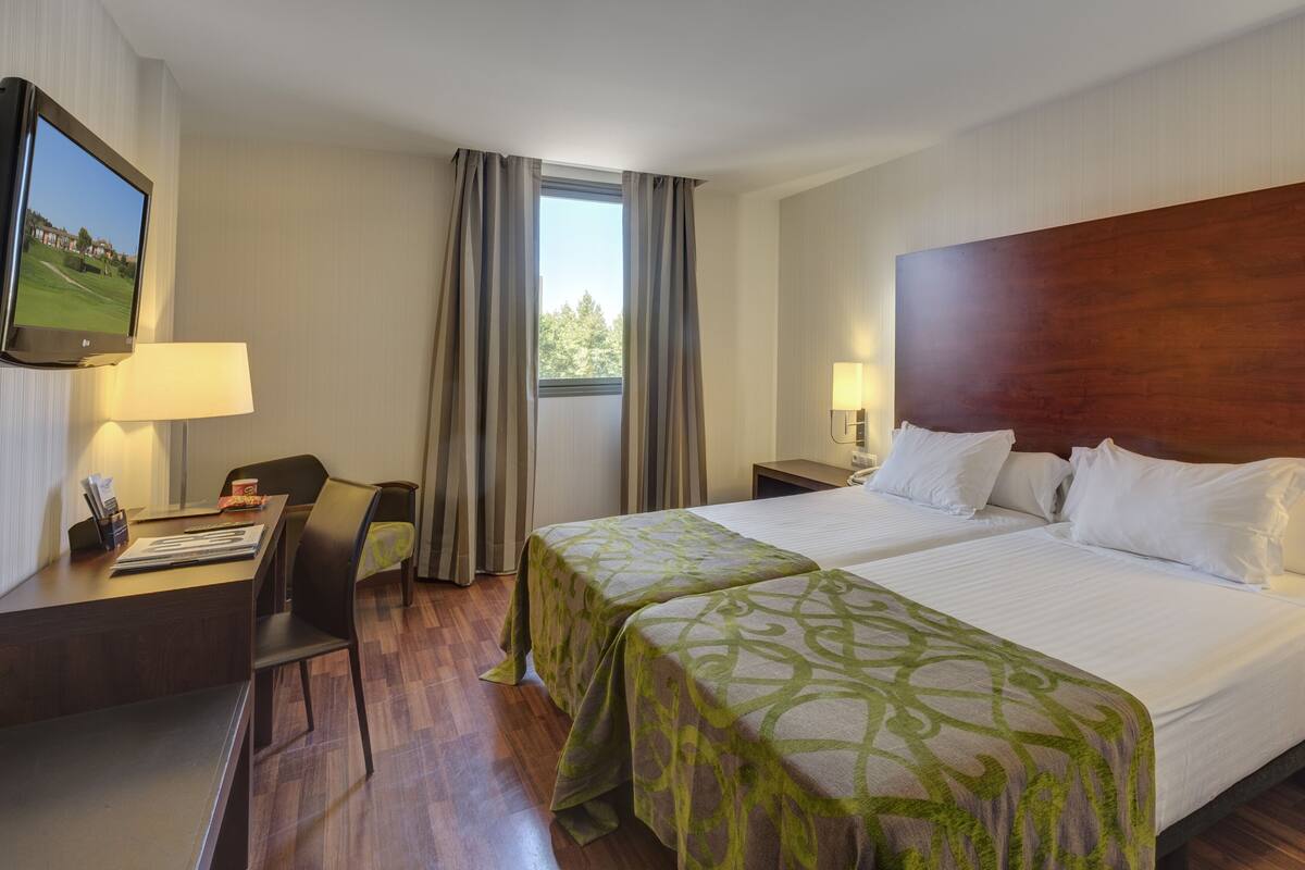 Ein Hotelzimmer mit zwei Betten, einem Schreibtisch, einem Stuhl, einem Fernseher und einem Fenster mit Vorhängen.
