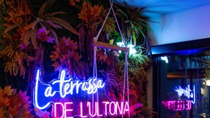 Un letrero de neón que dice "La terrassa de Lultrona" en una habitación con piso de madera y una silla colgante tipo hamaca.